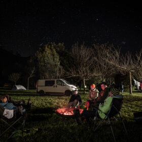 Nighttime camping scene with a group around a glowing campfire, van and tents under a starry sky.