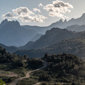 Winding dirt path through a forested mountain valley with jagged peaks in the distance under a partly cloudy sky.] , not; fix