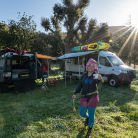 Smiling woman in a colorful outfit runs across a grassy campsite with two camper vans and kayaks on their roofs under a sunny sky.