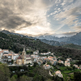 Hillside village with a tall church steeple, tan-roofed houses, and terraced greenery against distant snow-capped mountains under a cloudy sky.
