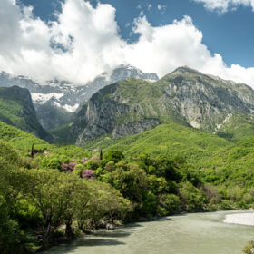 Lush alpine valley with a winding river, dense forest, and snow-capped peaks under a blue sky.