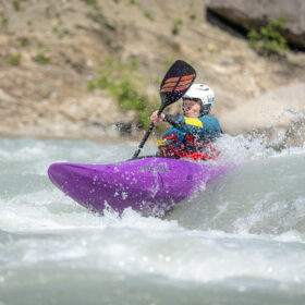 Kayaker wearing a white helmet paddling through whitewater in a purple kayak on a river rapids.