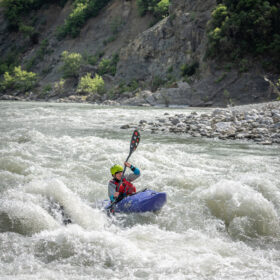 Kayaker wearing a helmet paddles through whitewater rapids in a blue kayak beside a rocky riverbank with a green forested hillside.