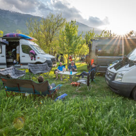 Group of people camping with parked campers and vans on a grassy field, mountains in the background.