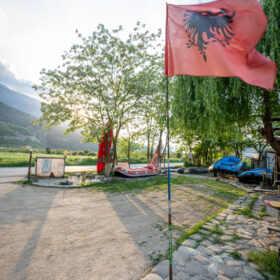Red Albanian flag with black double-headed eagle flutters on a pole near a gravel area with boats and trees in a sunny riverside setting.