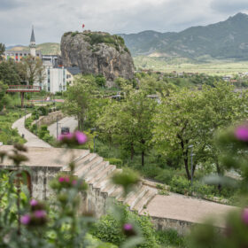 Park scene with a rocky hill and flag in the distance, surrounded by trees and buildings with mountains beyond