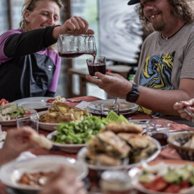 Two friends raise a glass as red wine is poured into a shared glass at a table filled with salads and plates of food.