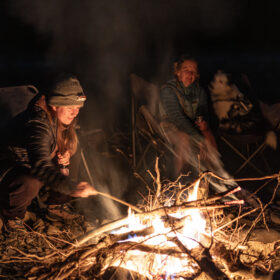 People sit around a campfire at night, roasting sticks; a person in a knit hat leans in as smoke rises between them.