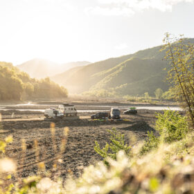 Sunlit riverbed with several vehicles parked and mountains in the background, under a bright sky.
