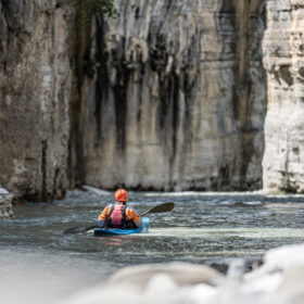 Person in orange helmet and life vest paddling a blue kayak through a rocky canyon river.