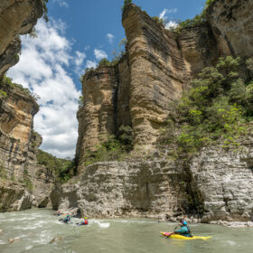 Kayakers paddling a rocky river between towering sandstone cliffs.