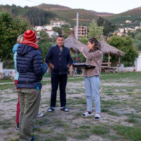 Outdoor group discussion: a woman with a clipboard speaks to several people in casual jackets, with thatched huts and hills in the background.