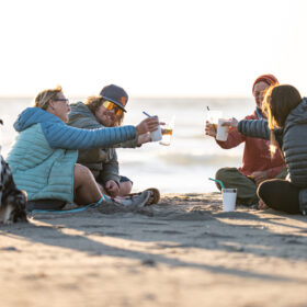 Friends on a sandy beach at sunset toast drinks together, with a dog sitting to the left.