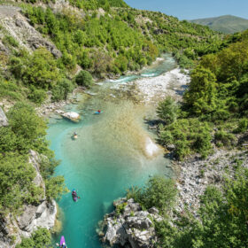 Teal river winds through a rocky, green canyon with kayakers paddling downstream under a clear blue sky.