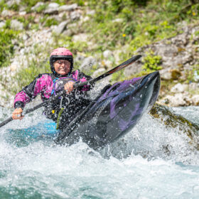 Kayaker wearing a pink helmet and jacket paddles a black kayak through whitewater rapids with spray around the bow.