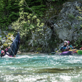 Person in pink helmet and blue life jacket in water beside a partially submerged kayak; three kayakers in colorful boats nearby.