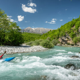 Kayaker in a red life jacket paddling a blue kayak on a fast, turquoise river with rocky banks and green trees.