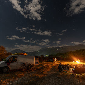 Night campsite: a white van with bikes attached, kayaks on the ground, and a group around a bright campfire under a starry sky near mountains.
