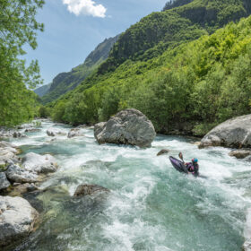 Rushing turquoise river through a rocky gorge with dense green trees and steep mountains in the background; a kayaker navigates rapids mid-channel.