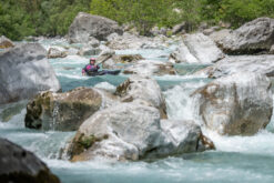 Kayaker wearing a pink helmet paddling through a rocky turquoise river. squinting water splashes around large boulders in a forested canyon.