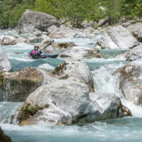 Kayaker wearing a pink helmet paddling through a rocky turquoise river. squinting water splashes around large boulders in a forested canyon.