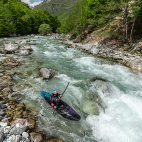 Kayaker wearing a pink helmet navigates whitewater in a rocky river canyon surrounded by green trees and distant mountains.
