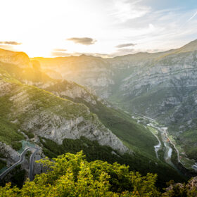 Sunlit mountain valley with winding road and river cutting through green cliffs at sunset.