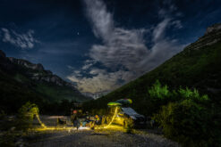 Nighttime campsite in a valley with string lights, a vehicle with a kayak on top, and people gathered around chairs in a rocky campsite.4a13a83d44d64f03aaelledEdit1