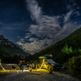 Nighttime campsite in a valley with string lights, a vehicle with a kayak on top, and people gathered around chairs in a rocky campsite.4a13a83d44d64f03aaelledEdit1