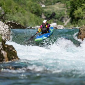 Kayaker wearing a helmet navigating whitewater in a blue-green kayak between rocks.