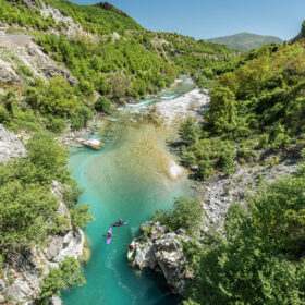 Turquoise river winding through a green forested canyon with kayakers paddling along rocky banks.
