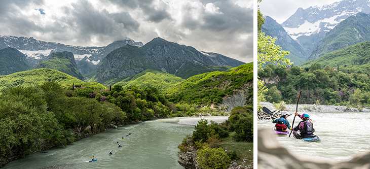 River winds through green mountains with snow-capped peaks in the distance; kayakers paddle downstream under a cloudy sky.