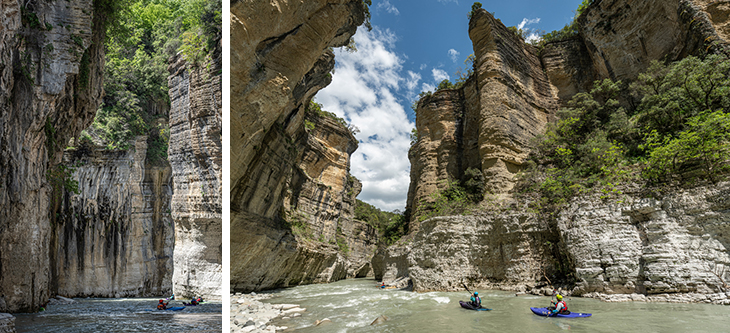 Two kayakers navigate a narrow river between towering limestone cliffs with green foliage above the canyon walls.