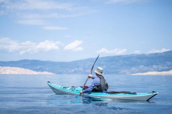 Person paddling a blue and white kayak on a calm lake, wearing a life vest and hat, with distant hills and blue sky behind him/her.