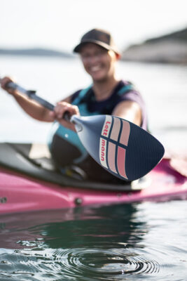Person in a pink kayak reaches the paddle toward the camera on calm water, smiling as they paddle.