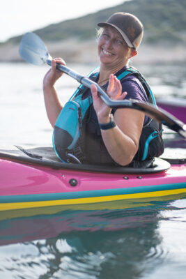 Smiling woman paddling a pink kayak, wearing a blue life jacket and cap on calm water.