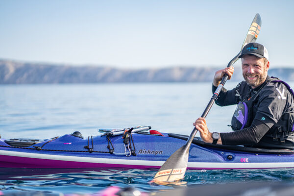 Man paddling a blue kayak on calm water, smiling, wearing cap and life vest with a paddle raised mid-stroke.