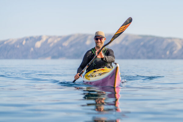 Man wearing a hat and sunglasses paddling a bright pink and yellow kayak on calm blue water with distant hills in the background,