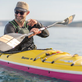 Smiling man in a hat and sunglasses paddles a bright yellow kayak on calm water, adjusting his paddle mid-stroke.