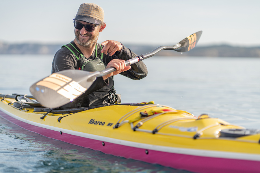 Smiling man in a hat and sunglasses paddles a bright yellow kayak on calm water, adjusting his paddle mid-stroke.