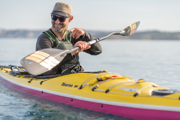 Smiling man wearing a hat and sunglasses paddling a yellow kayak on calm water.