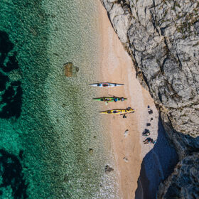 Aerial view of a sandy beach with four colorful kayaks lined up, turquoise water on the left and rocky cliffs on the right