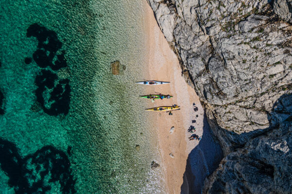 Aerial view of a sandy beach with four colorful kayaks lined up, turquoise water on the left and rocky cliffs on the right