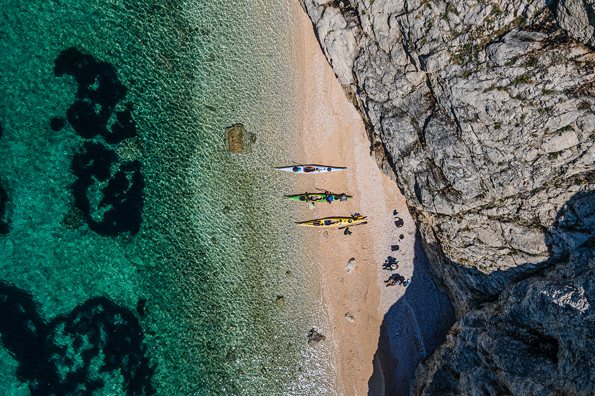 Aerial view of a sandy beach with four colorful kayaks lined up, turquoise water on the left and rocky cliffs on the right