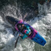 Kayaker in a teal jacket and turquoise helmet paddles a bright blue-pink kayak through white-water rapids.