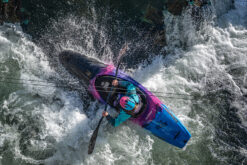 Kayaker in a teal jacket and turquoise helmet paddles a bright blue-pink kayak through white-water rapids.