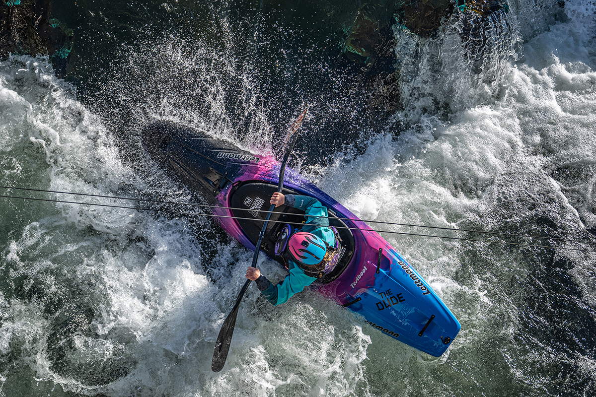 Kayaker in a teal jacket and turquoise helmet paddles a bright blue-pink kayak through white-water rapids.