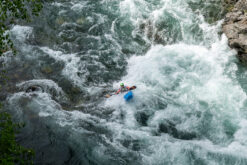 Kayaker wearing a green helmet paddling through rough white-water rapids in a blue kayak