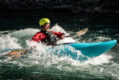 Kayaker wearing a yellow helmet and red jacket paddles a turquoise kayak through whitewater rapids.