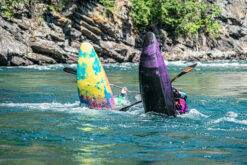 Two brightly colored paddle boards capsized in a fast-flowing turquoise river near a rocky, forested shore, with a paddler in a wetsuit nearby reaching for a paddle.